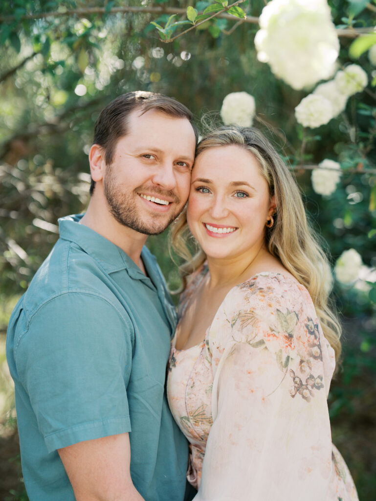 couple smiling cheek to cheek surrounded by florals for their engagement session