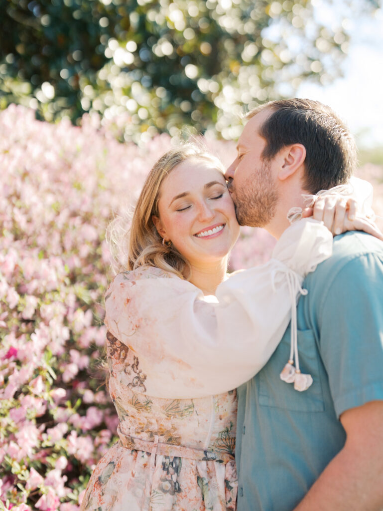 man kissing woman on cheek surrounded by pink florals Dreamy Outdoor Engagement Photos in Botanical Gardens