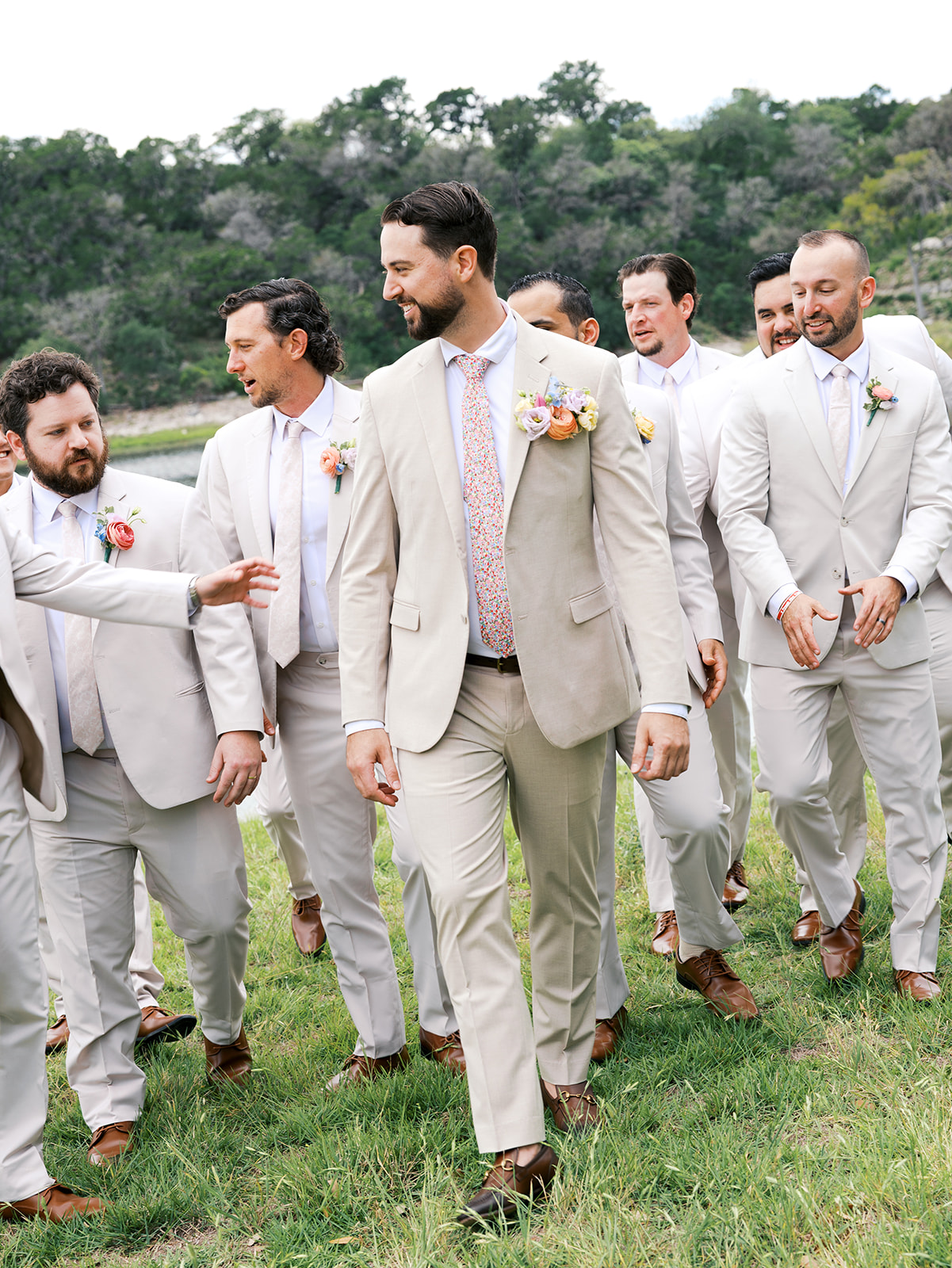 A group of men in light-colored suits with boutonnieres walk together outdoors on grass, with one man in front leading the group for a wedding in Austin, TX