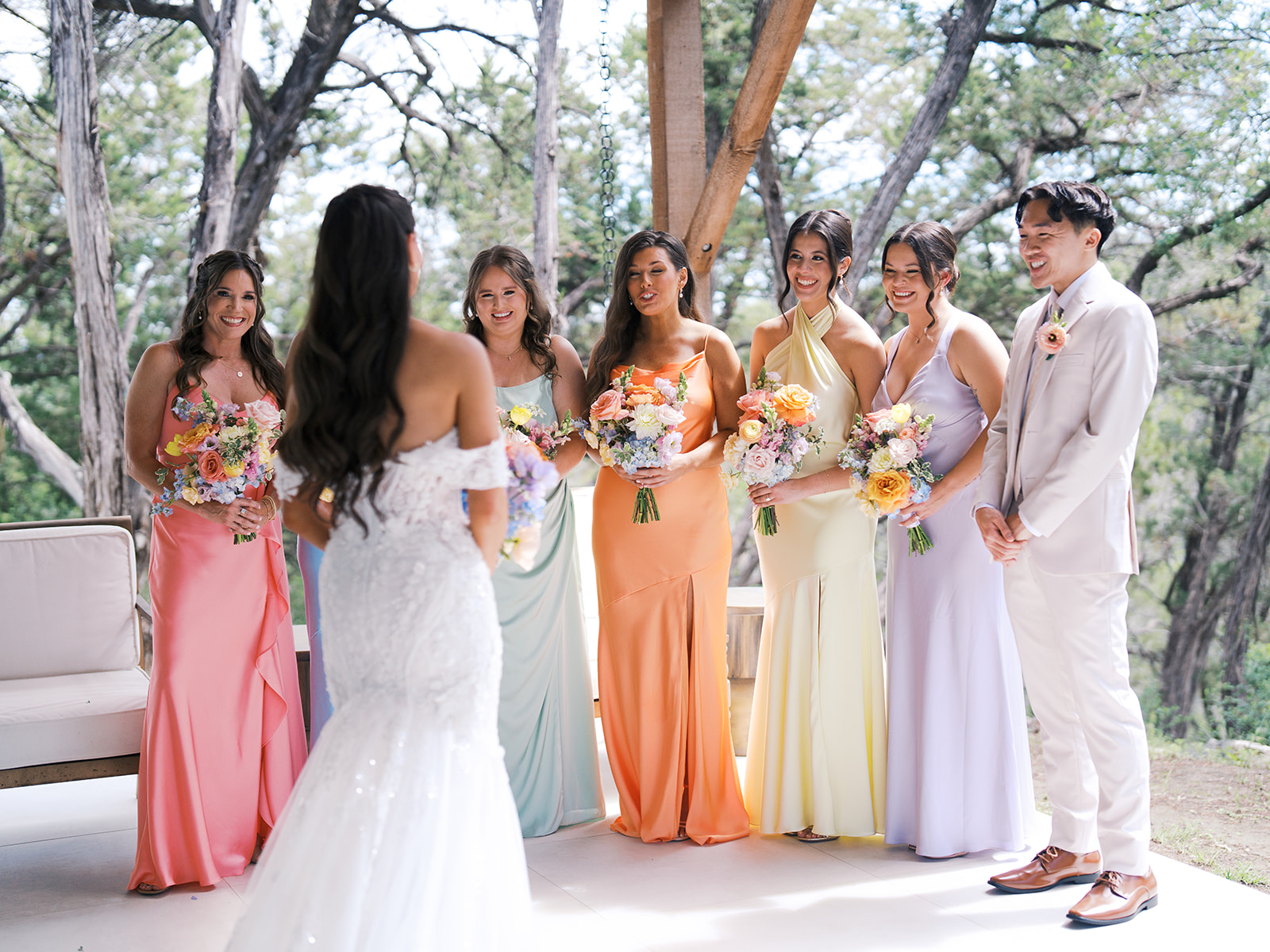 A bride in a white dress faces five bridesmaids in pastel gowns and a groomsman in a light suit, all holding bouquets, standing outdoors.
