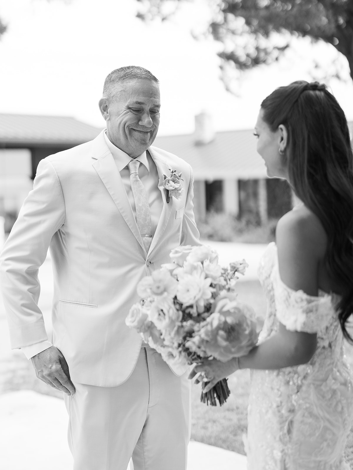 A man in a light suit smiles at a woman in a wedding dress holding a bouquet outdoors.