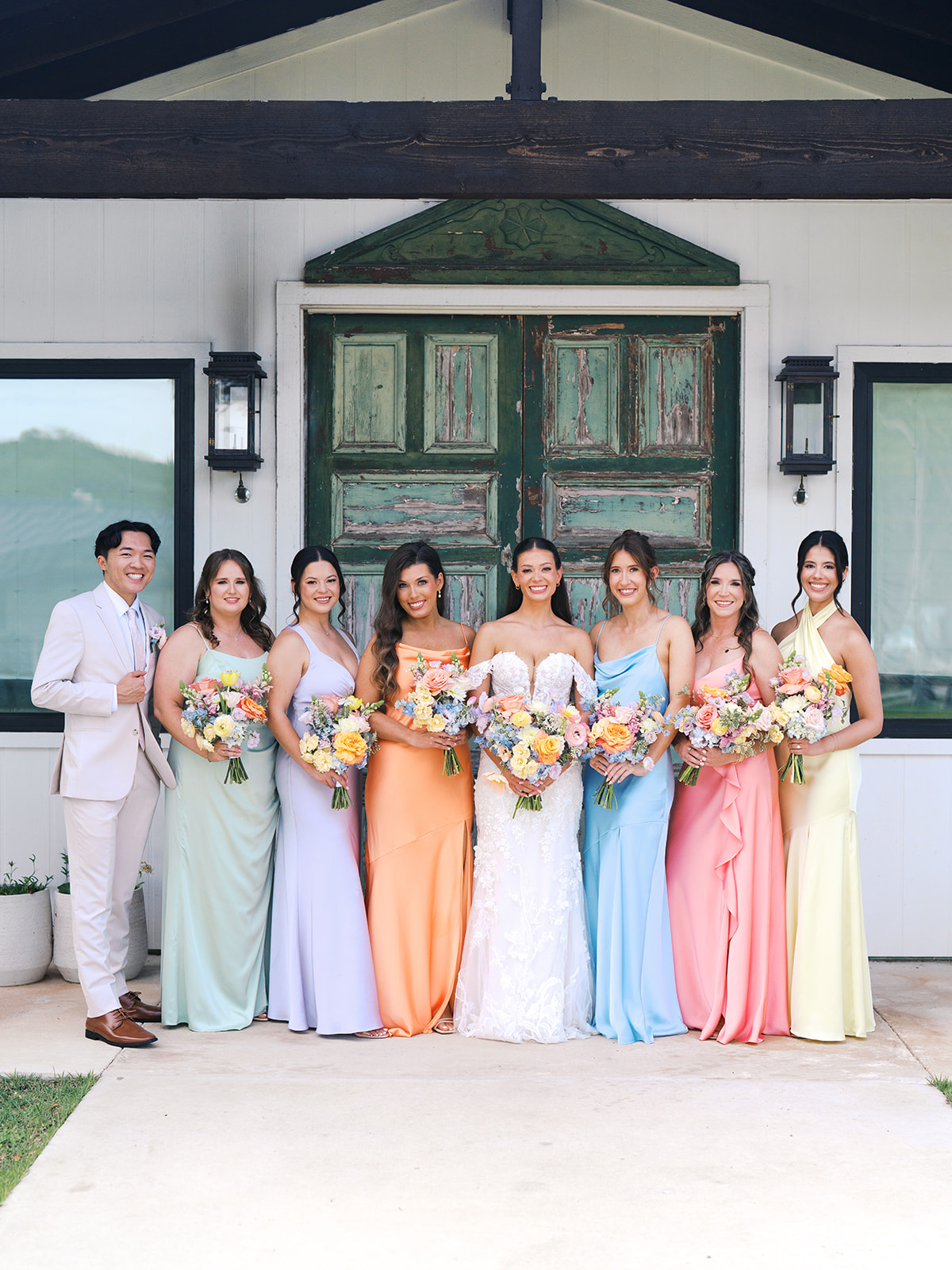 A bride in a white dress stands with seven bridesmaids in colorful dresses and one man in a light suit, all holding bouquets, posing in front of a rustic green door for a wedding in austin tx