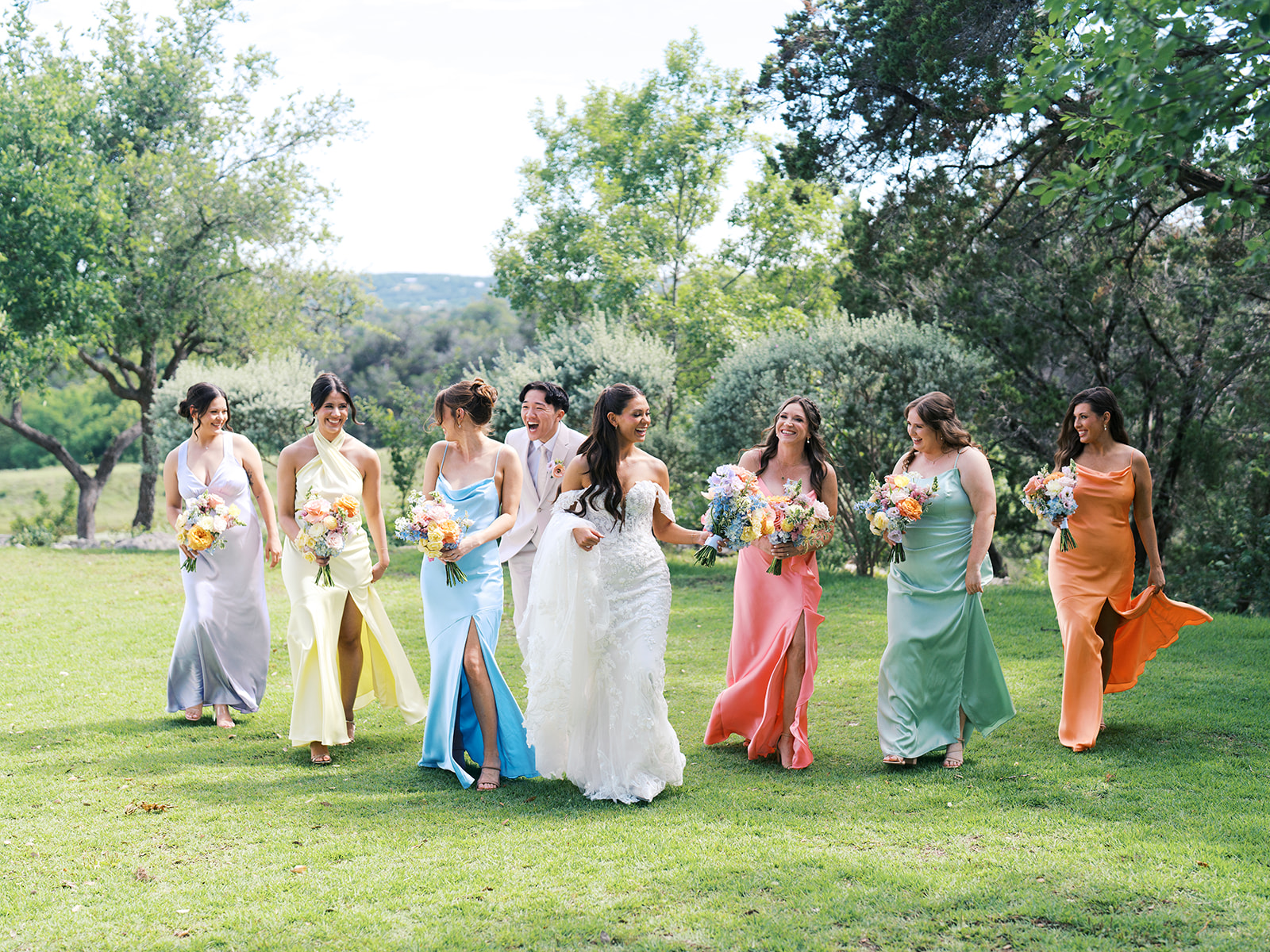 A bride and seven bridesmaids in colorful dresses walk together on grass, holding bouquets, with trees in the background.