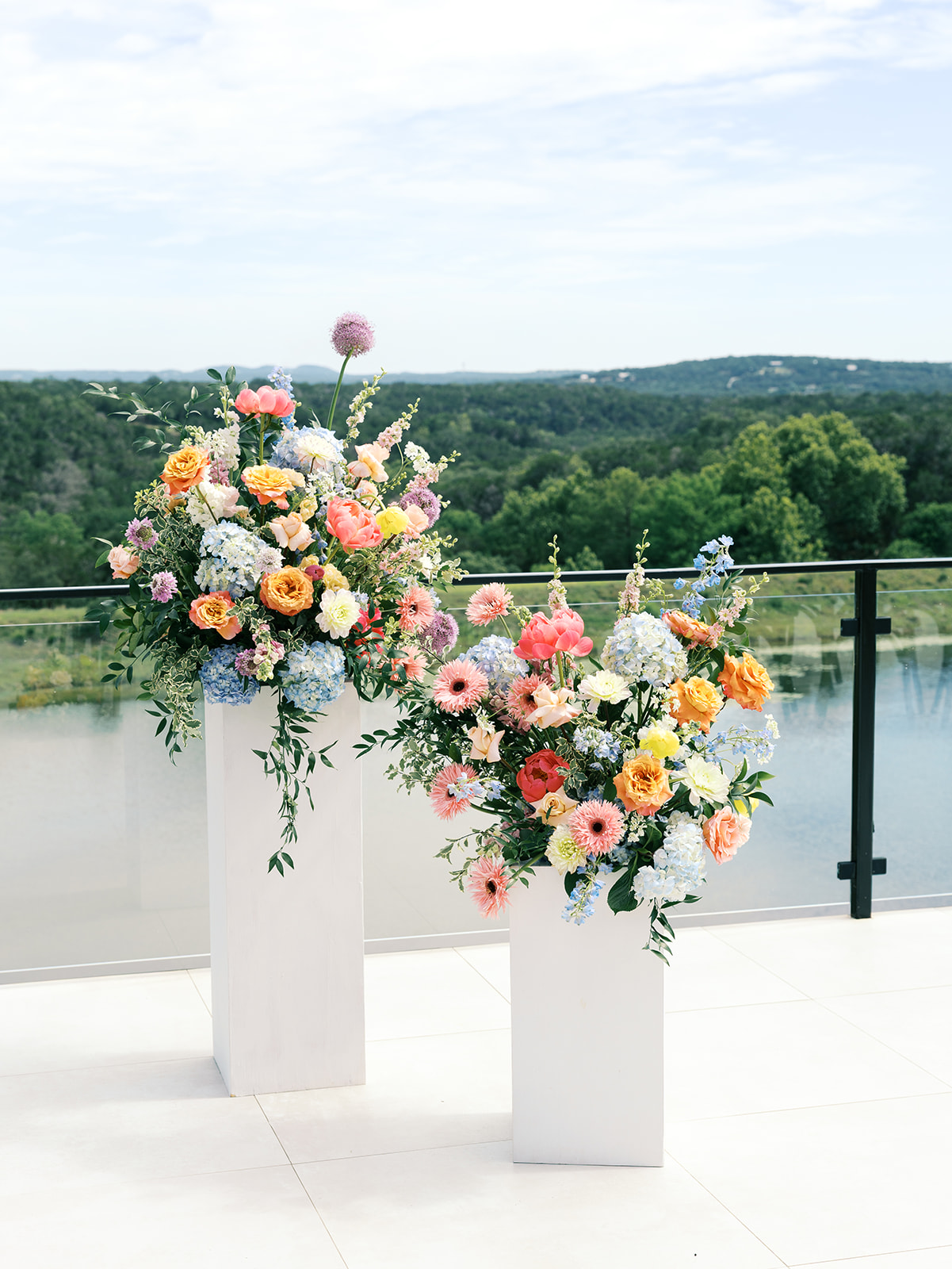 Two white pedestal stands hold large, colorful floral arrangements on a patio with a glass railing overlooking a river and green landscape for a wedding in austin tx