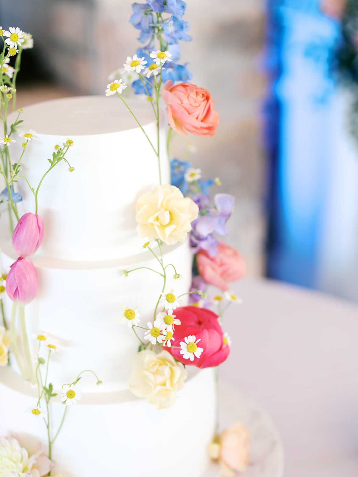 A three-tiered white cake decorated with colorful fresh flowers, including pink, yellow, and purple blooms, displayed on a round table.