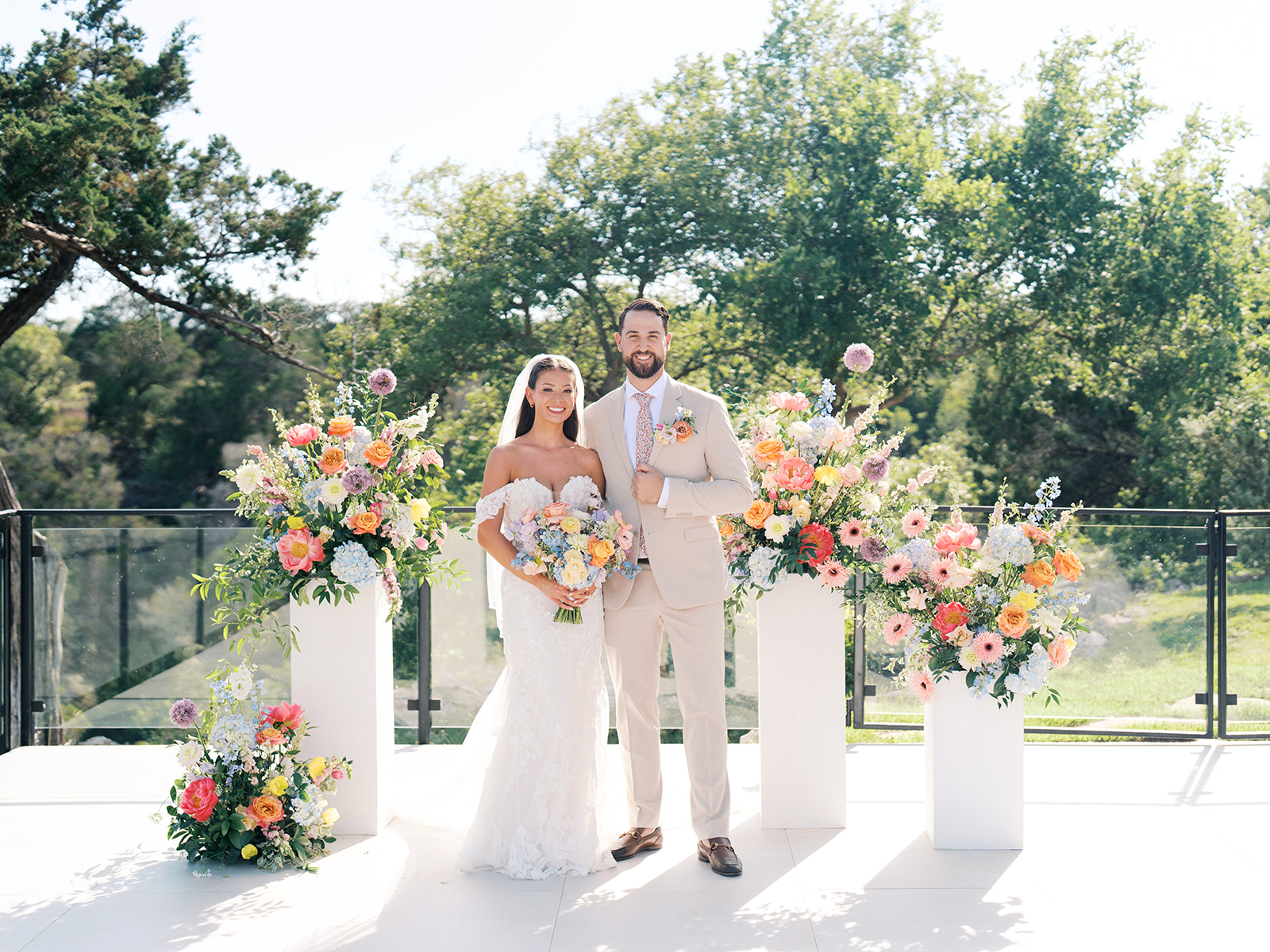 A bride and groom stand together outdoors, surrounded by large floral arrangements on a sunny day