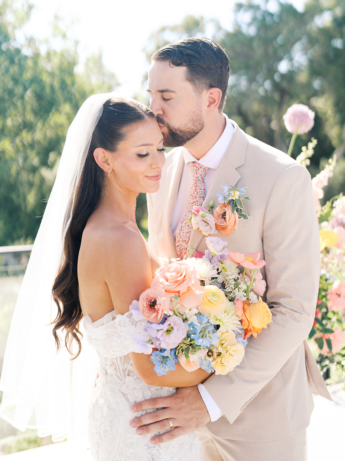 Bride and groom embrace outdoors; groom kisses bride's forehead while holding a colorful bouquet for a wedding in austin tx