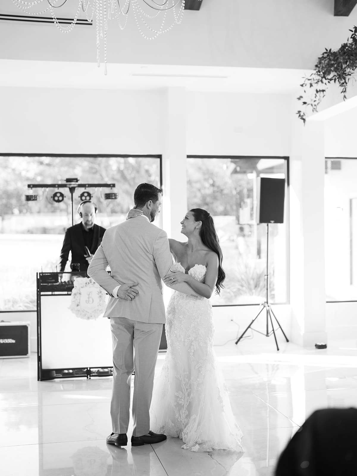 A bride and groom dance together in a bright, modern venue while a DJ stands behind a booth in the background.