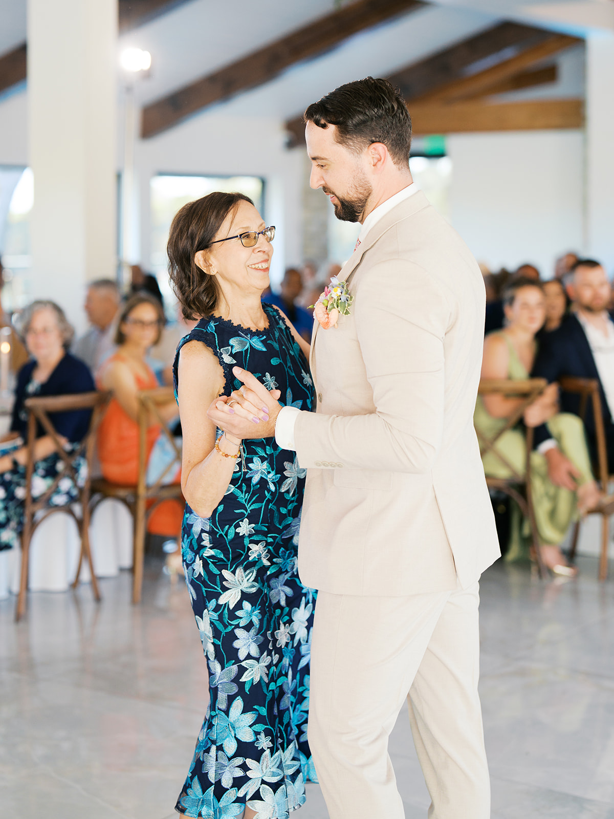 A man in a light-colored suit dances with a woman in a blue floral dress at an indoor event, with seated guests watching in the background.
