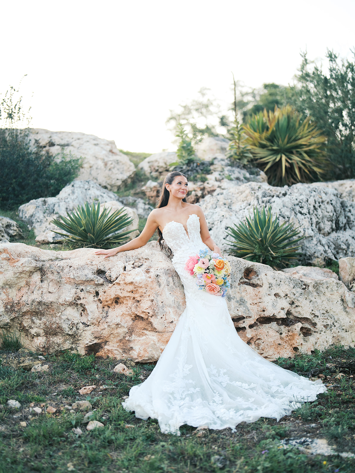 A bride in a strapless white wedding gown stands outdoors against large rocks, holding a colorful bouquet of flowers for a wedding in austin tx