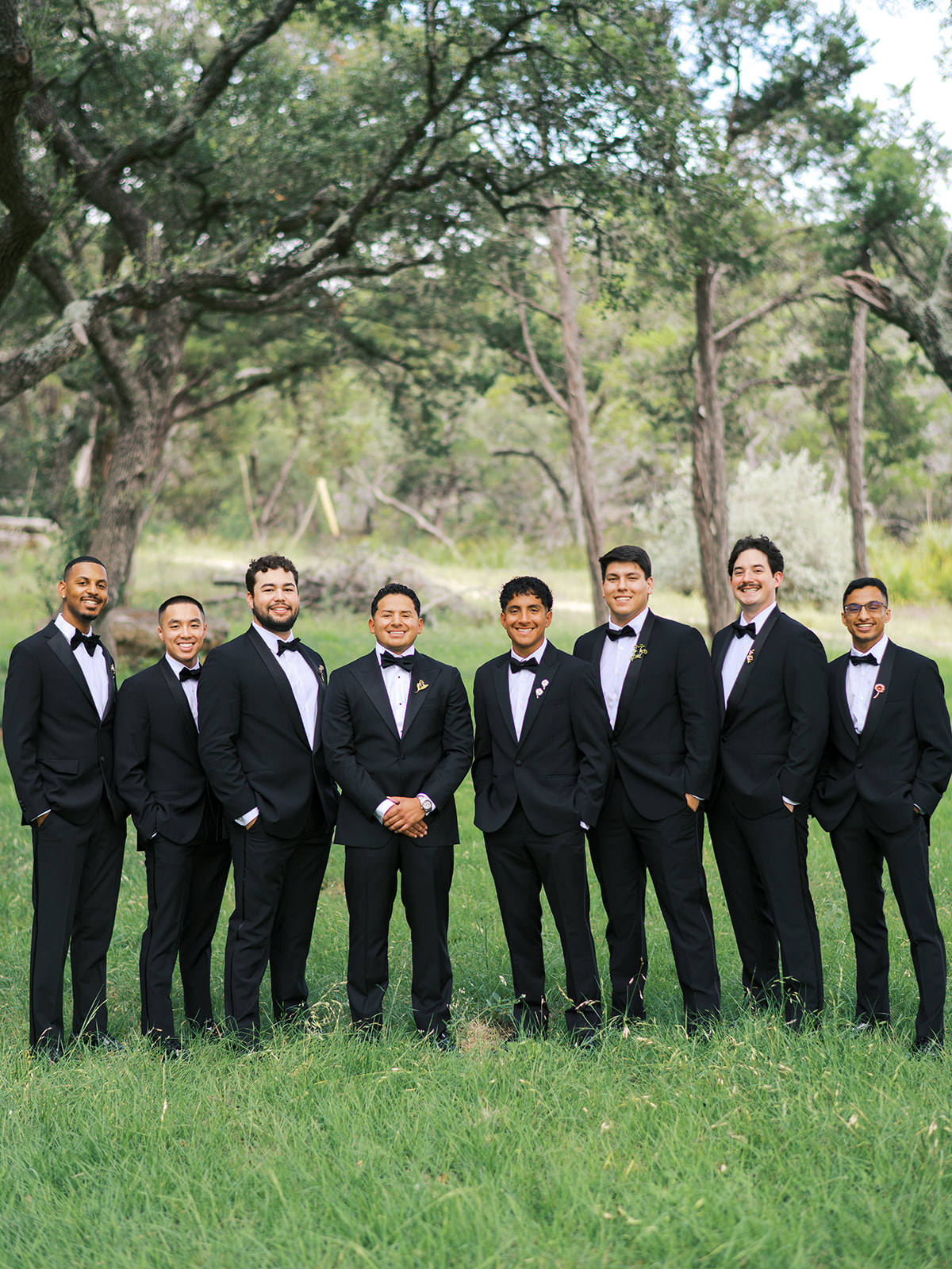 Eight men in black tuxedos stand in a row on grass with trees in the background, posing for a formal group photo outdoors.
