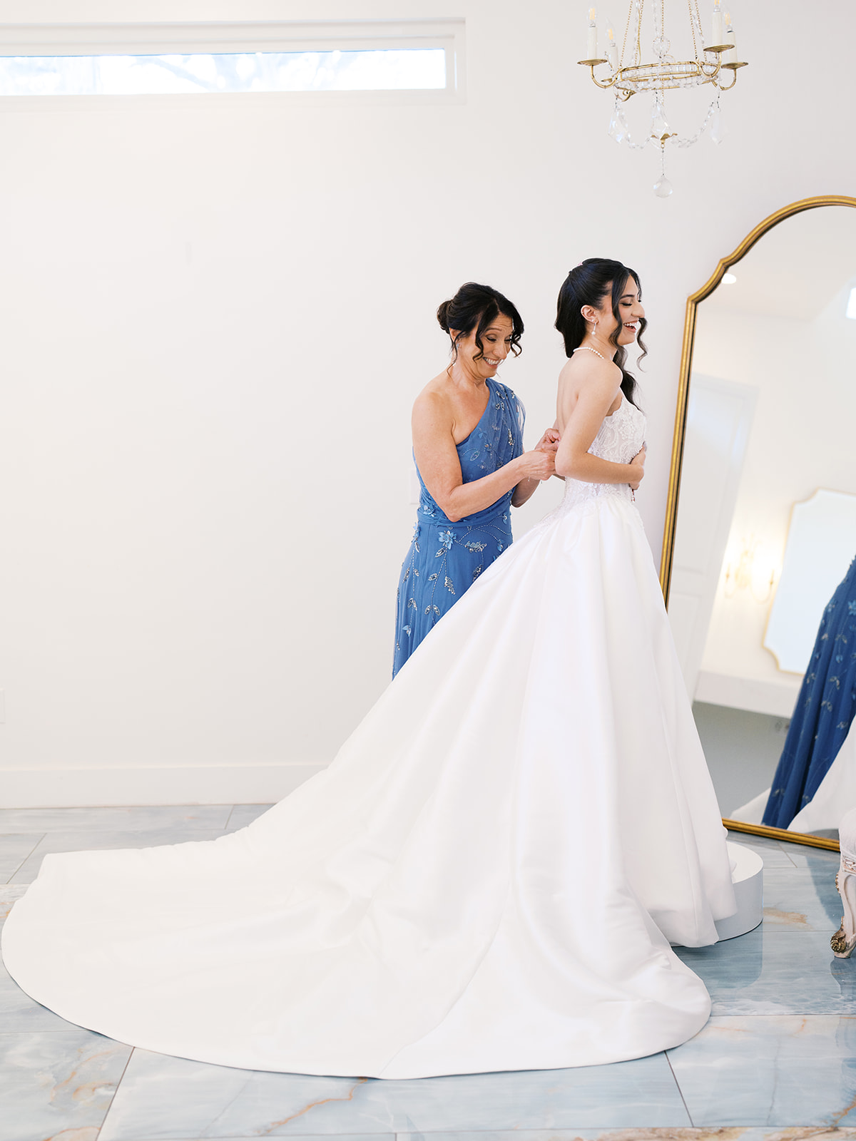 A woman in a blue dress helps another woman in a white wedding gown adjust her dress in a bright room with a large mirror for a wedding at the videre estate 