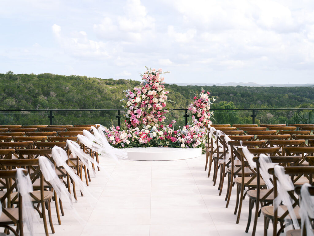 Rows of wooden chairs line an outdoor wedding aisle with white fabric accents, leading to a floral altar arrangement against a green landscape backdrop fora wedding at the videre estate 
