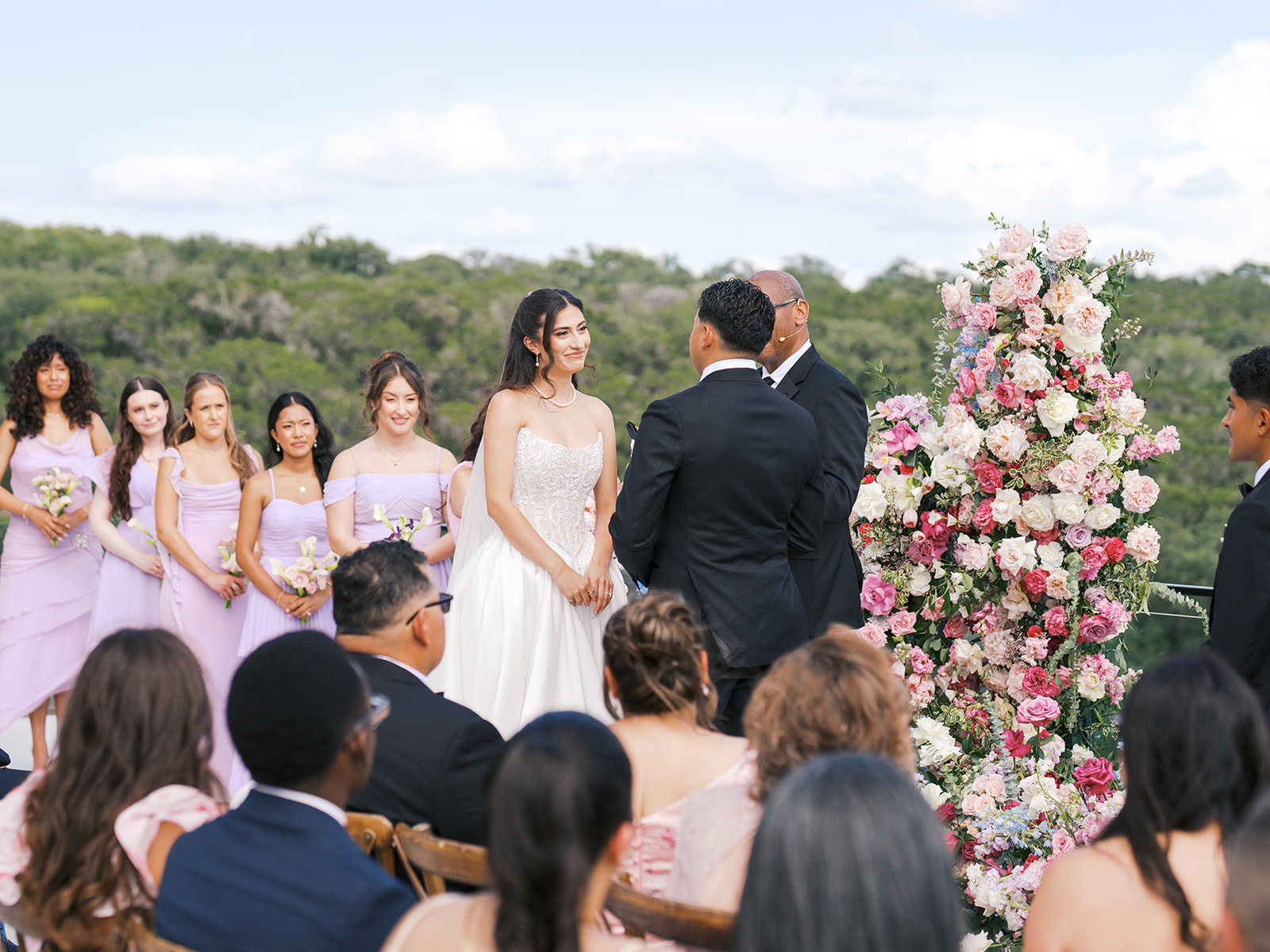 A bride and groom stand at an outdoor altar with bridesmaids, groomsmen, and guests seated, surrounded by greenery under a partly cloudy sky for a wedding at the videre estate