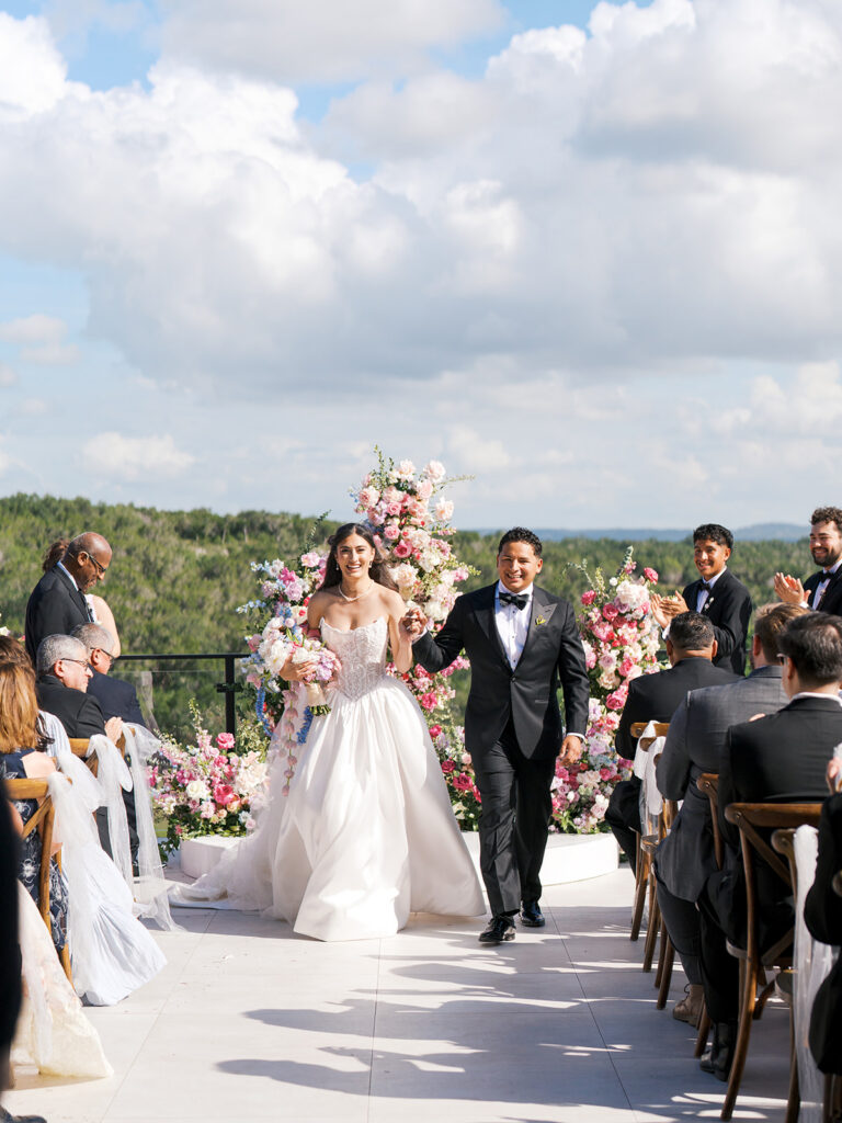 A bride and groom walk down the aisle outdoors, surrounded by guests and pink floral arrangements, under a partly cloudy sky.