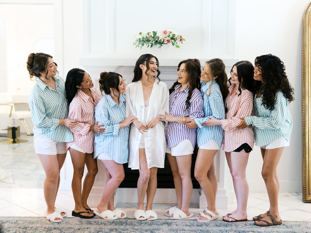 Seven women in matching striped shirts and shorts stand indoors, gathered around a woman in a white robe, all smiling and facing each other in a bright, elegant room for a wedding at the videre estate 
