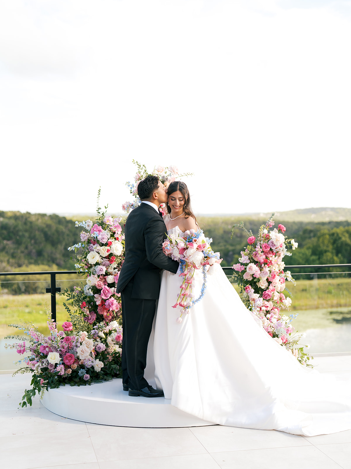 A bride and groom stand on a white platform outdoors, surrounded by pink and white floral arrangements, with a scenic natural background at a Texas hill country wedding at The Videre Estate