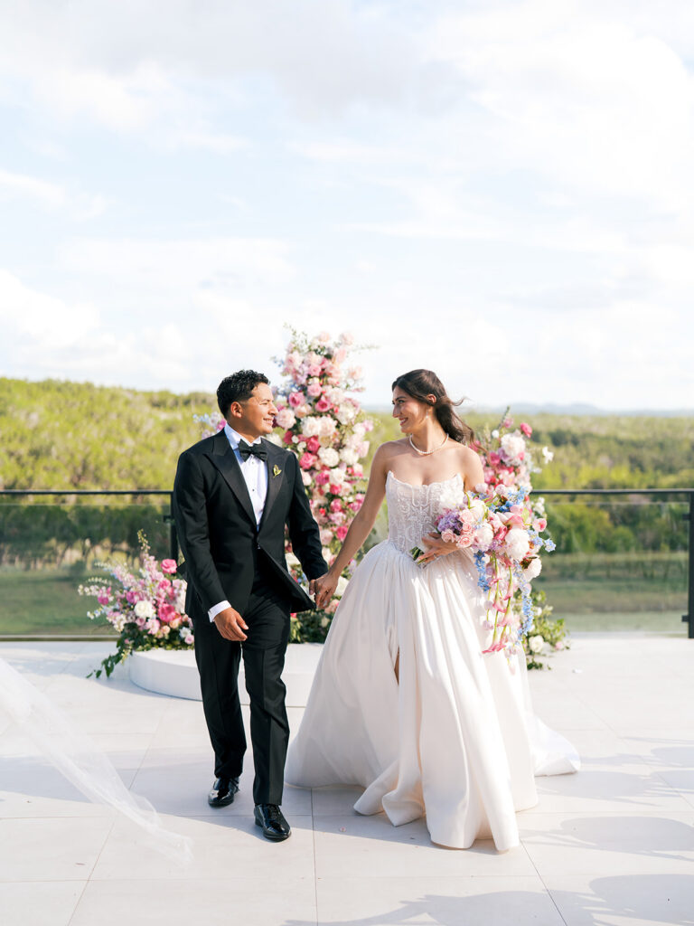 A bride and groom walk down the aisle outdoors, surrounded by guests and pink floral arrangements, under a partly cloudy sky.