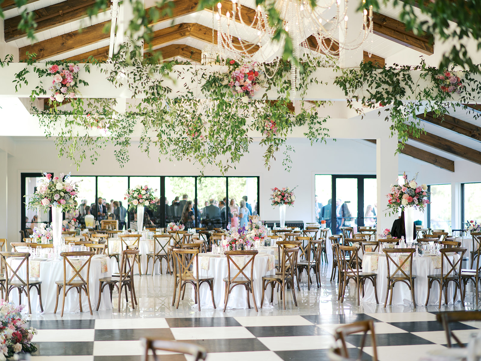 A spacious wedding reception hall with wooden chairs, round tables with white linens, floral centerpieces, hanging greenery, and large windows.