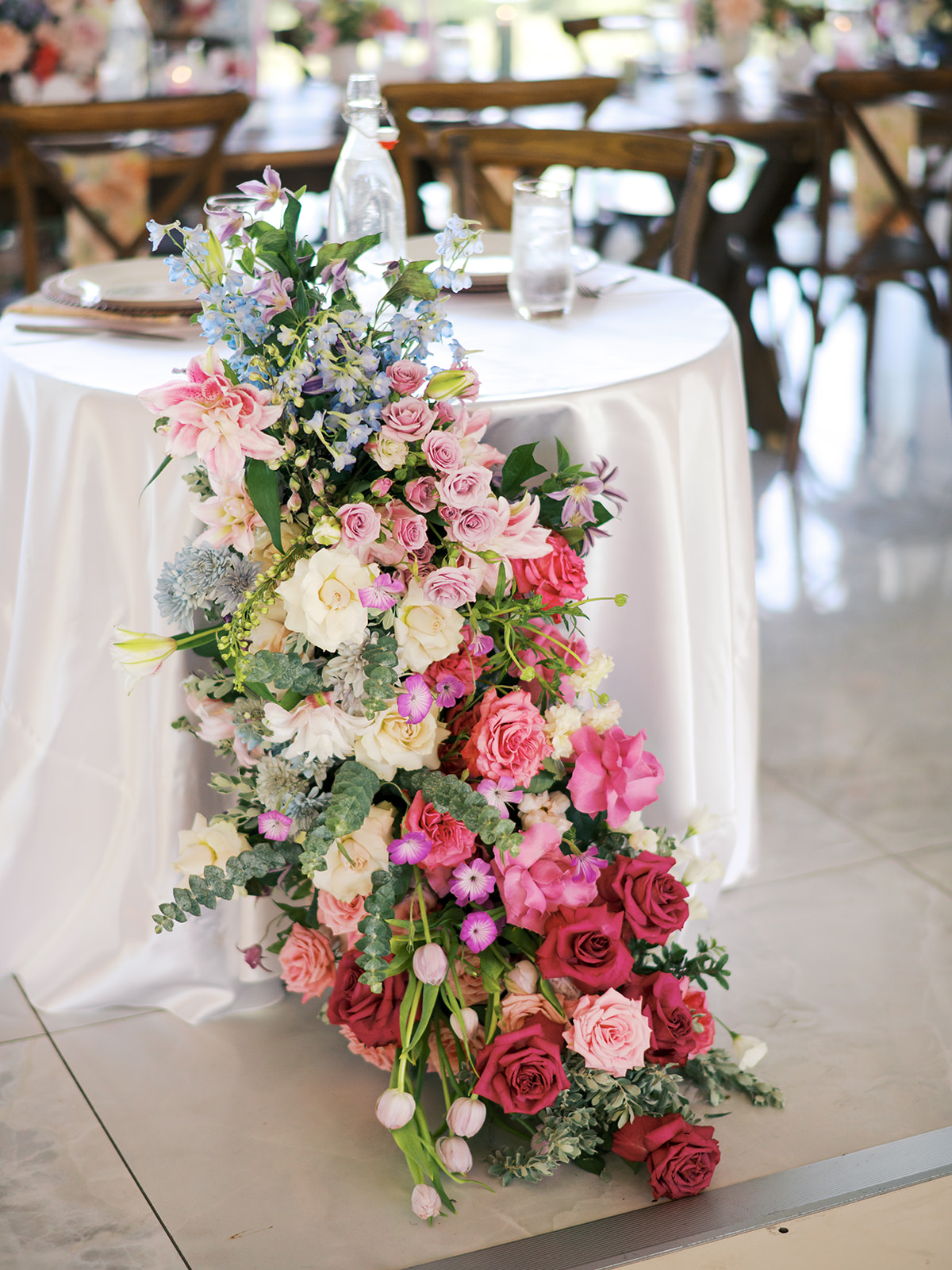 A wooden table is set with plates, glassware, pastel candles, and a large floral centerpiece featuring pink, white, and purple flowers for a wedding at the Videre Estate