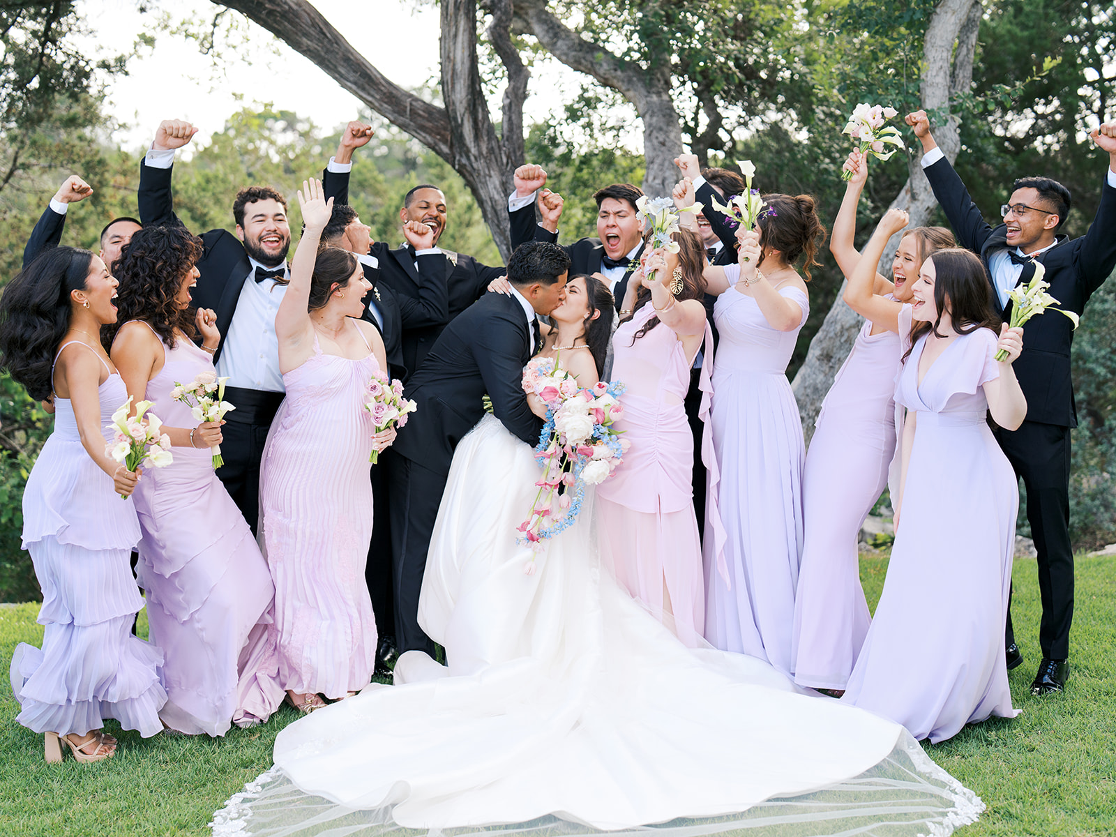 A bride and groom kiss outdoors, surrounded by a cheering wedding party dressed in black suits and lavender dresses for a wedding at the videre estate