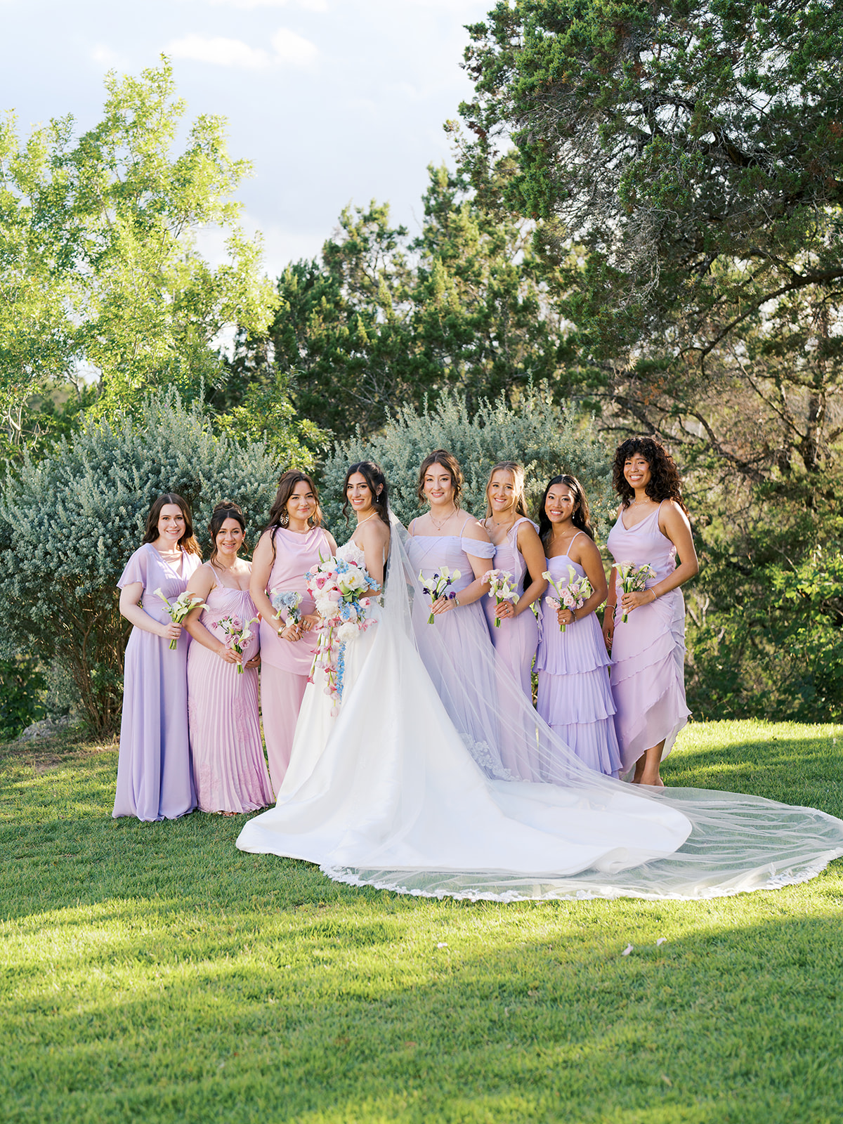A bride in a white gown stands outdoors with six bridesmaids wearing pastel dresses, all holding bouquets, on a grassy area with trees in the background.