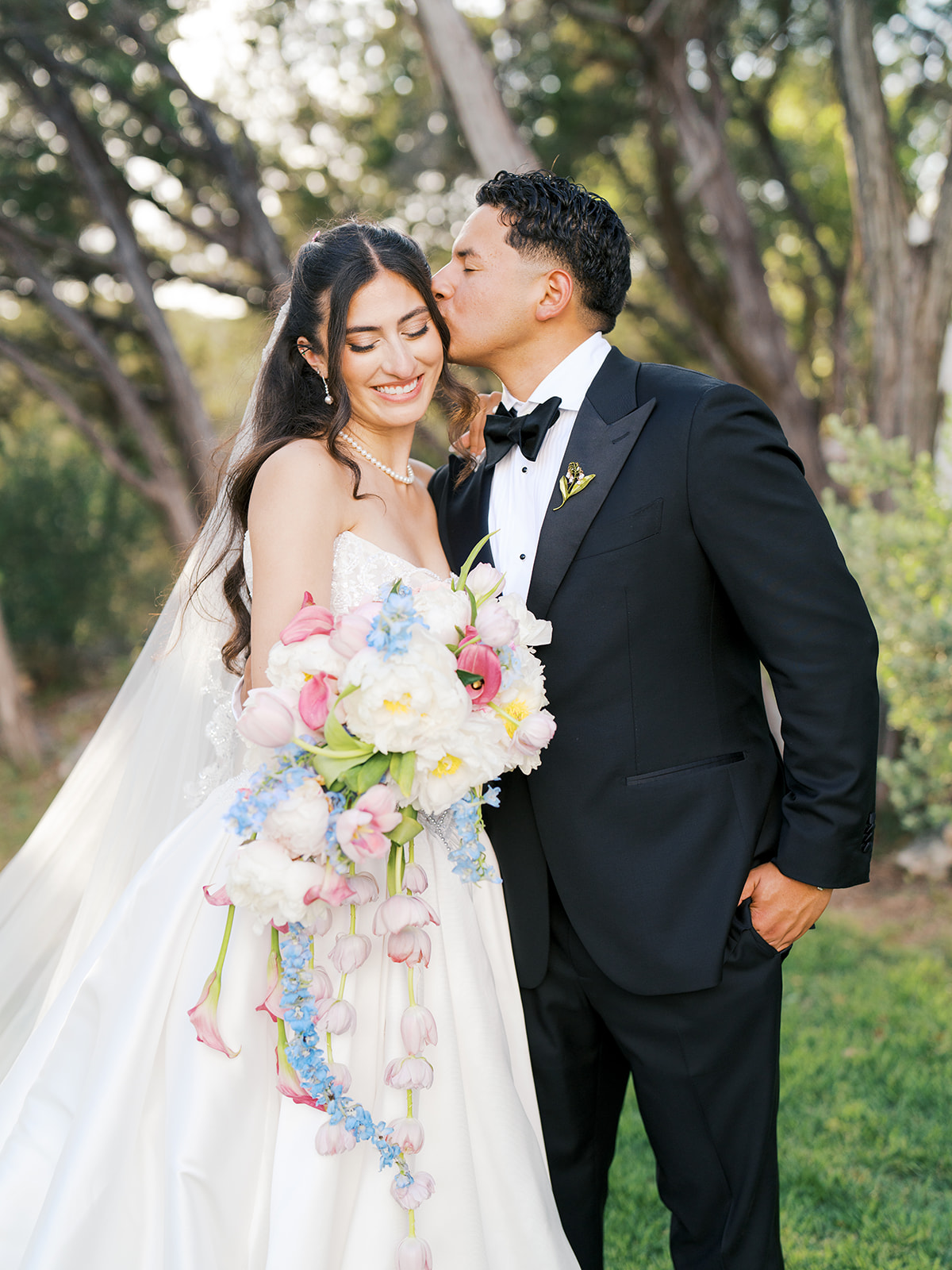 A groom in a black tuxedo kisses the bride on the temple as she smiles, holding a colorful bouquet outdoors for a wedding at the videre estate 