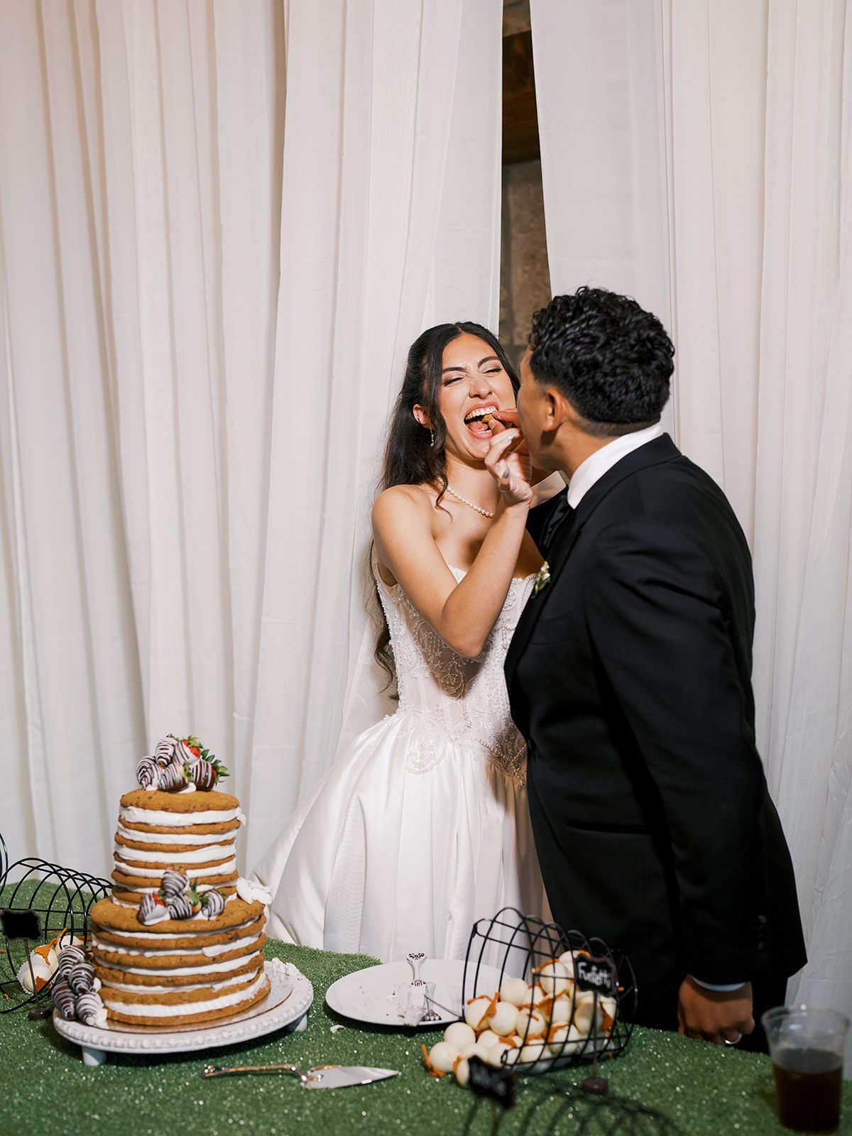 A bride and groom in formal attire cut a tiered wedding cake together 
