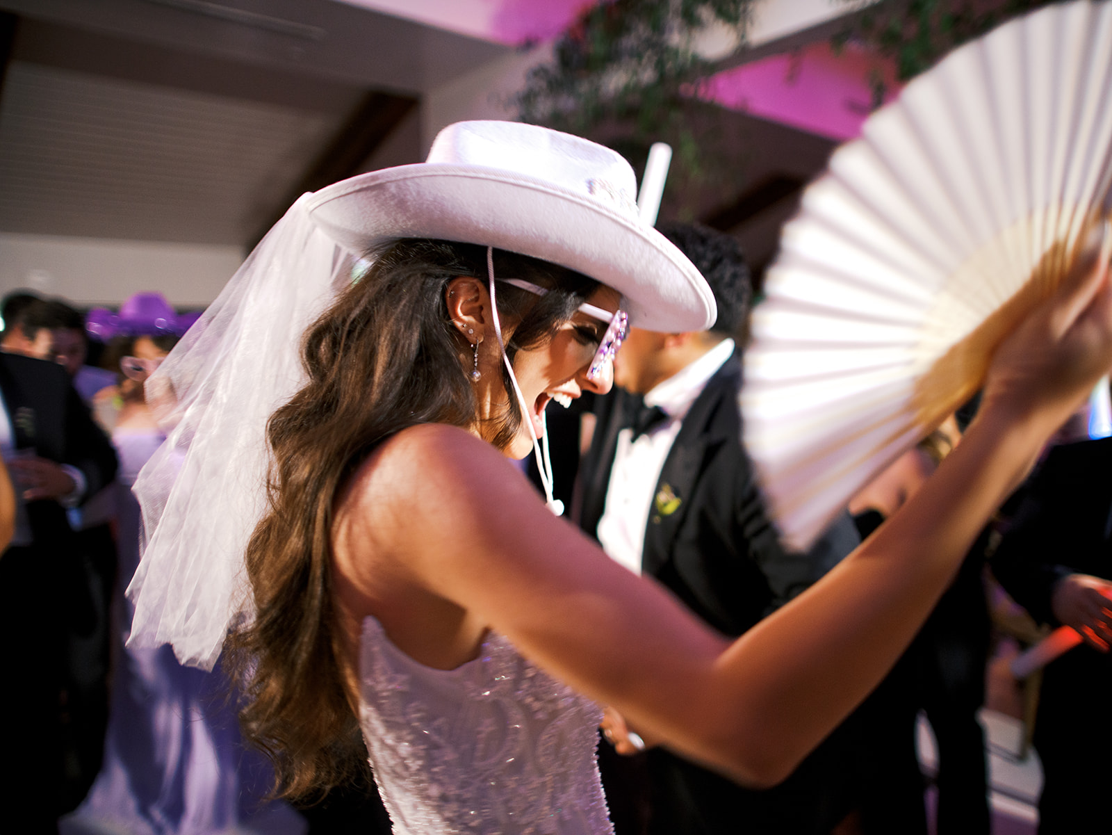 A woman in a wedding dress and cowboy hat dances with a white fan among a group of people at a lively indoor event.