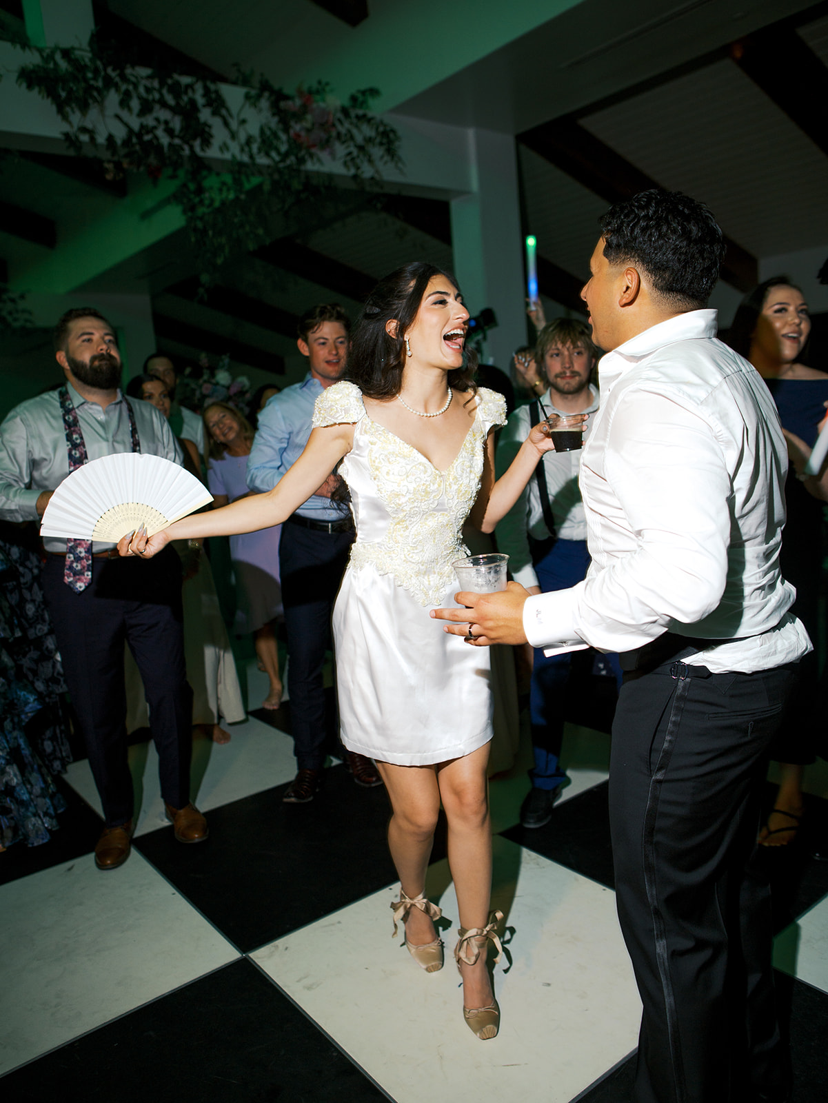 A woman in a short white dress and a man in a white shirt dance together at a party, surrounded by other people on a black and white checkered floor.