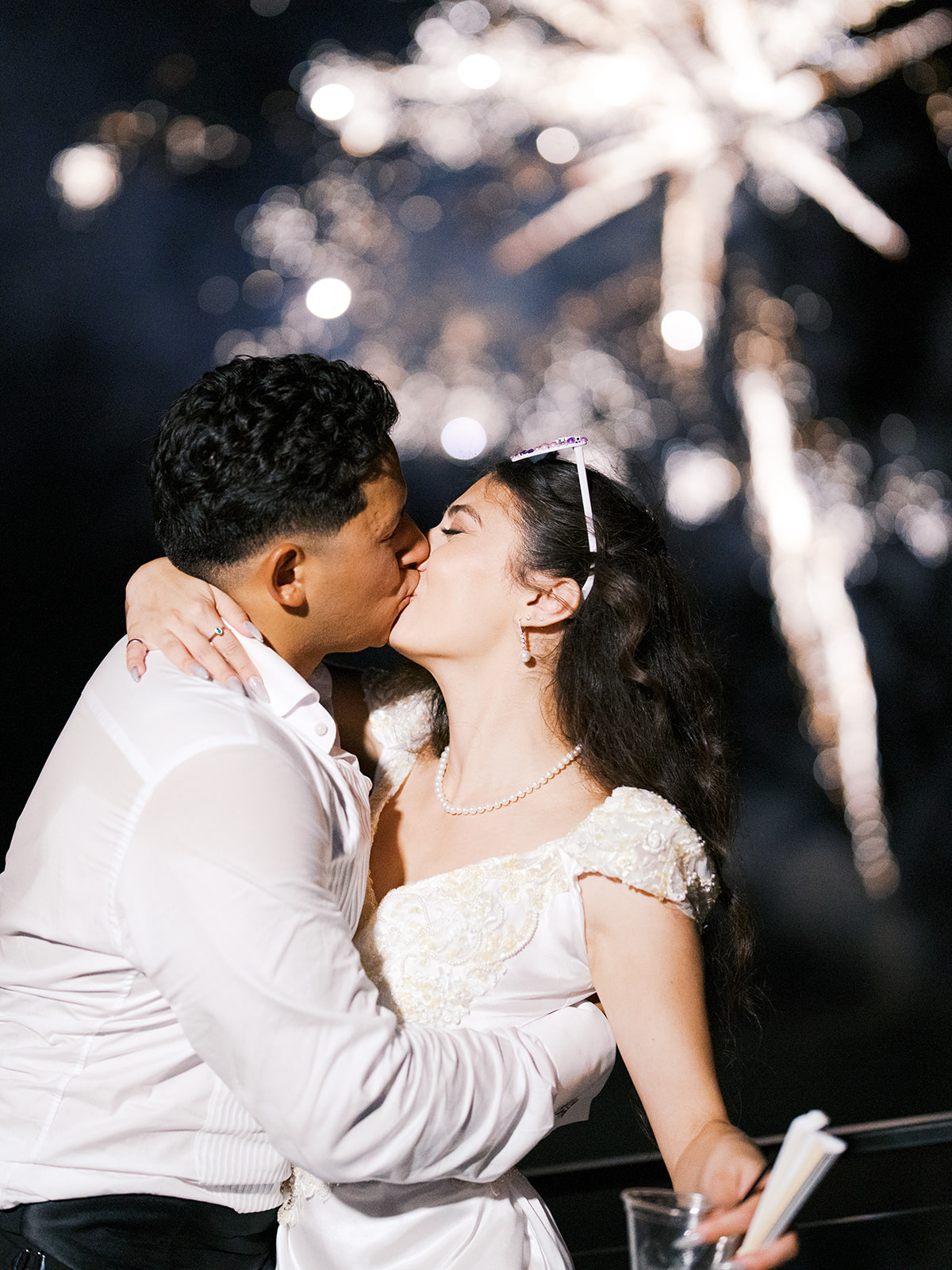 A couple dressed in white kiss passionately at night with fireworks exploding in the sky behind them.