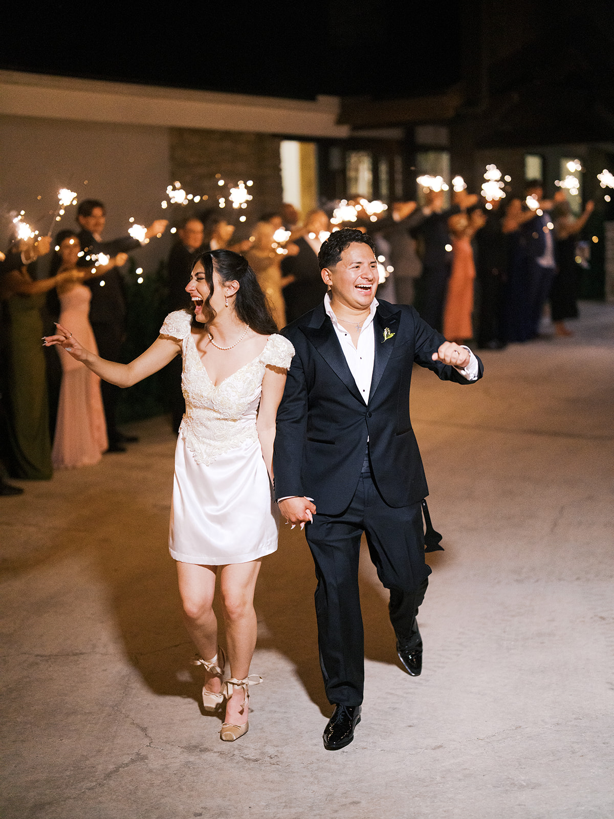 A couple walks hand in hand outdoors at night, smiling, while guests in the background hold sparklers.
