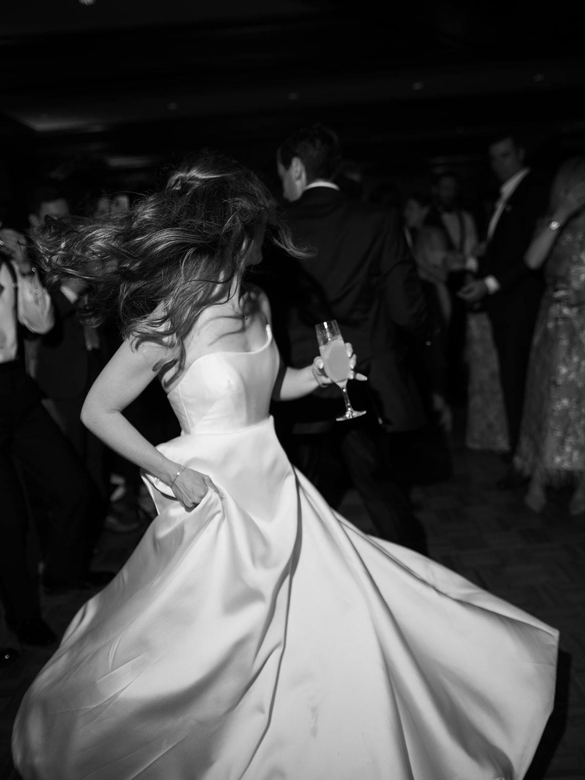 A woman in a strapless gown holding a drink twirls on a crowded dance floor at a formal event.