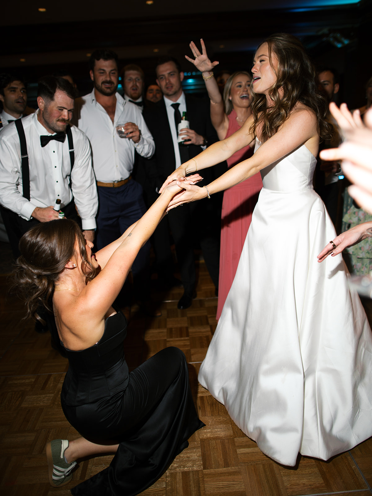 A bride in a white dress holds hands with a woman in a black dress kneeling on the dance floor, surrounded by people in formal attire at a lively party.