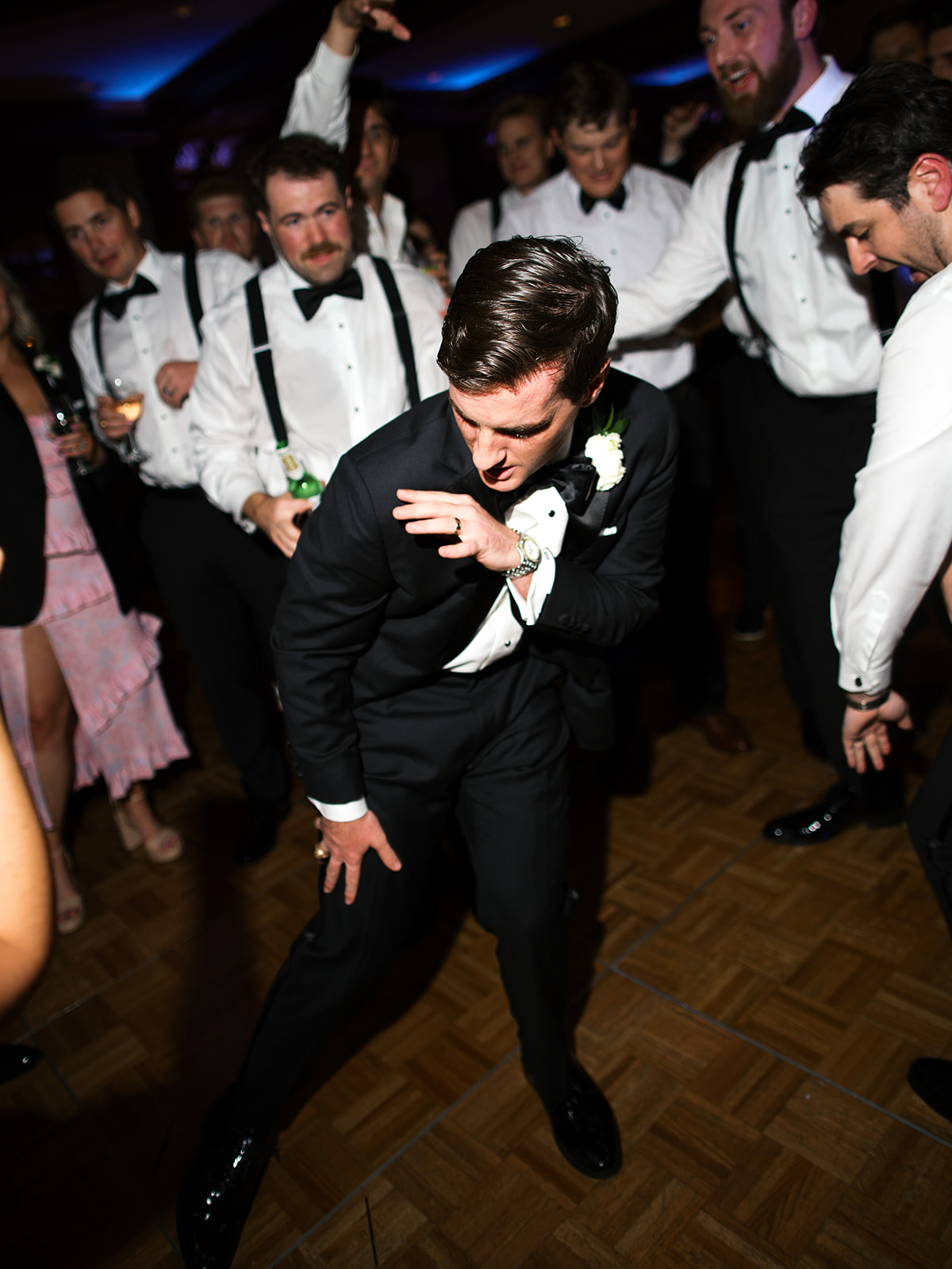 A man in a suit dances energetically on a wooden floor, surrounded by people in formal attire at a lively indoor event for a Houstonian Hotel Wedding