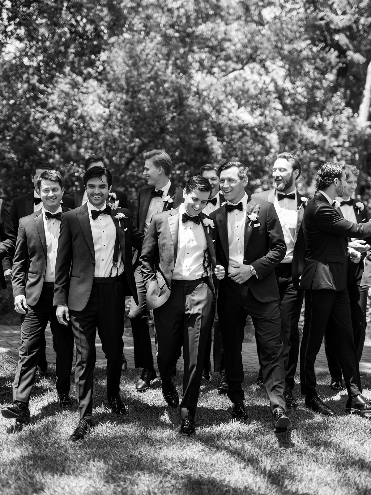 A group of men in tuxedos walk together outdoors on grass, with trees in the background. The scene appears to be at a formal event, such as a wedding.