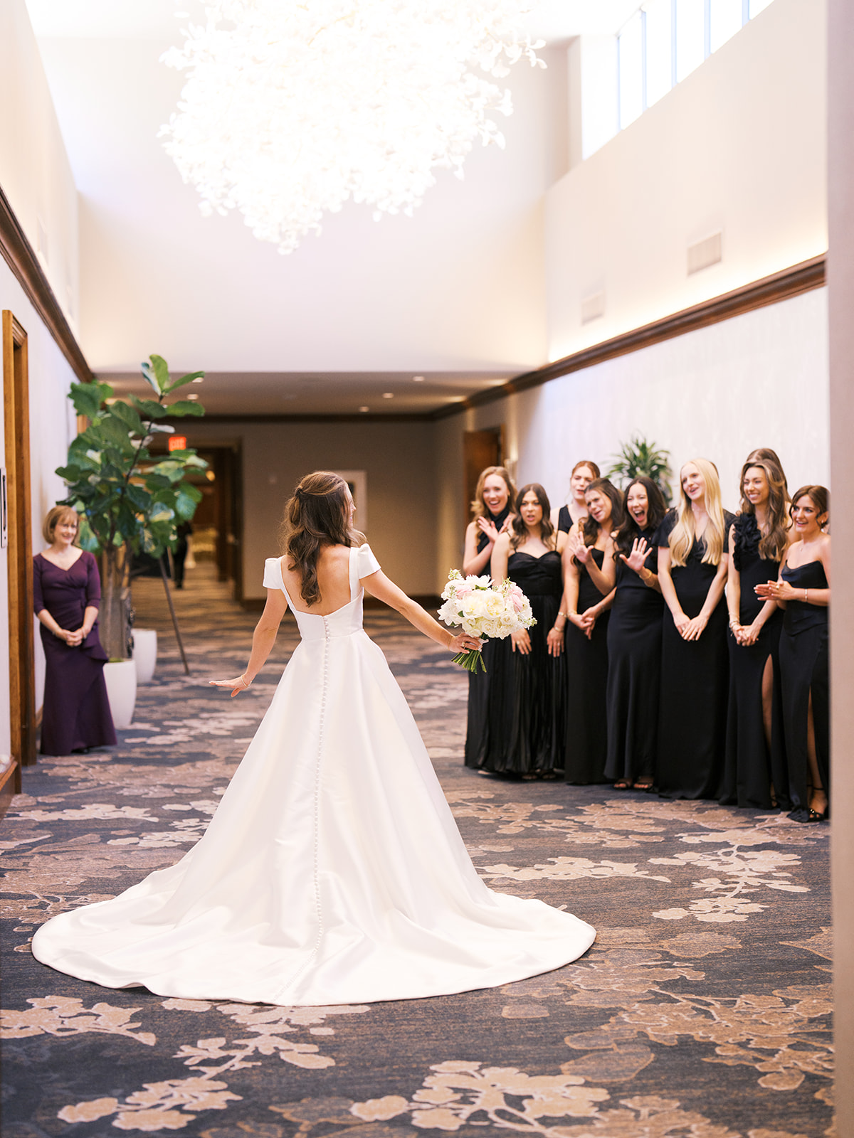 A bride in a white gown stands with her back to the camera, holding a bouquet, as a group of bridesmaids in black dresses react with excitement in a hallway for a Houstonian Hotel Wedding