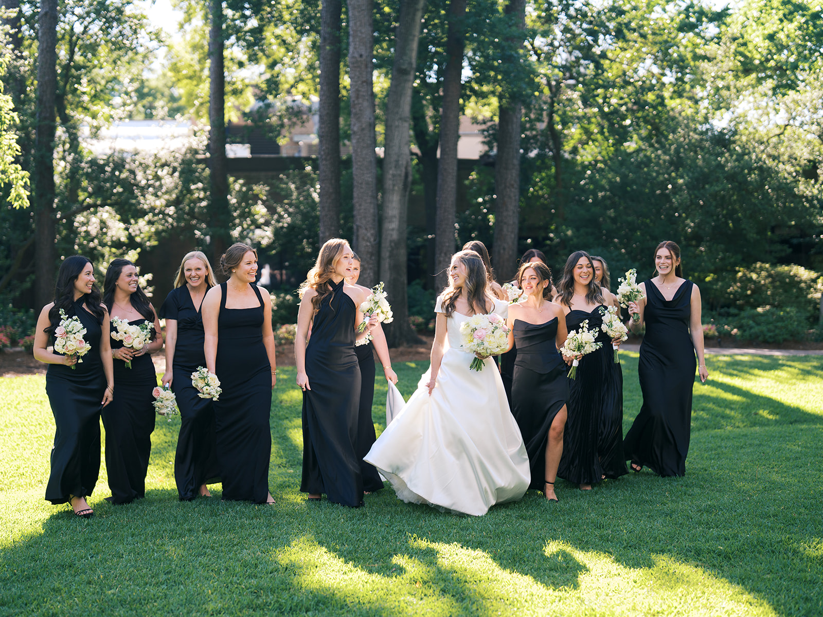 A bride in a white dress walks on grass with seven bridesmaids in black dresses, all holding bouquets, under tall trees in a sunlit outdoor setting for a Houstonian Hotel Wedding