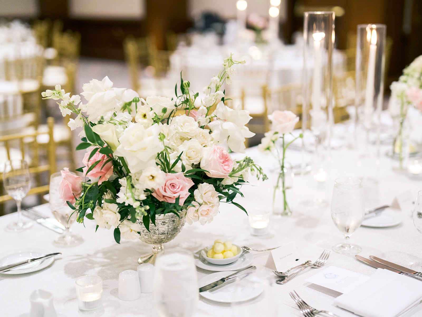 Elegant floral centerpiece with white and pink flowers on a round table set for a formal event, featuring white tablecloth, glassware, candles, and gold chairs in the background.