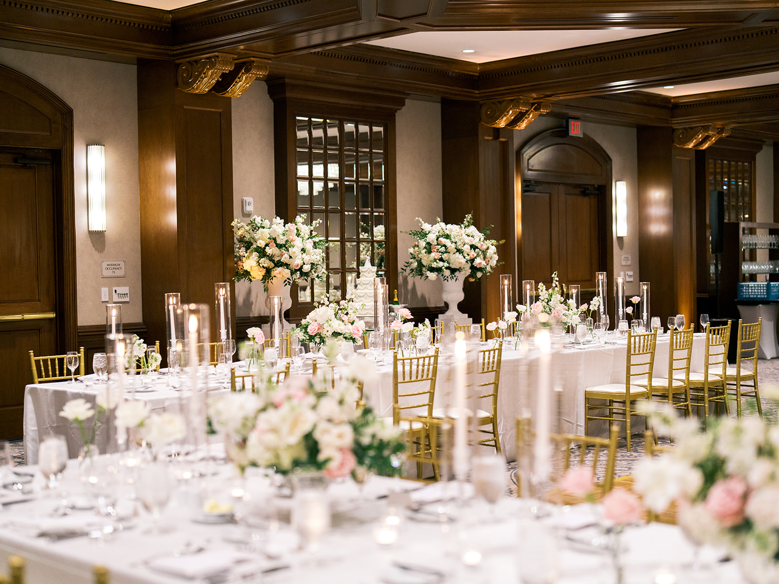 Elegant floral centerpiece with white and pink flowers on a round table set for a formal event, featuring white tablecloth, glassware, candles, and gold chairs in the background.