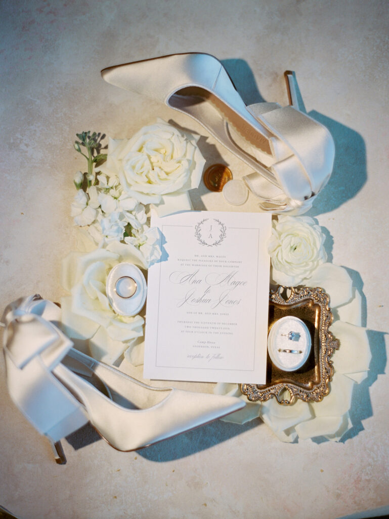 Flat lay of white high heels, wedding invitation, white roses, candles, and a framed ring box arranged on a light surface.