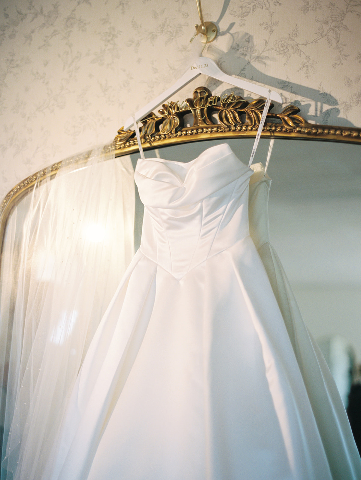 A white satin wedding dress with a full skirt hangs on a hanger in front of an ornate gold-framed mirror, with a veil draped to the side.