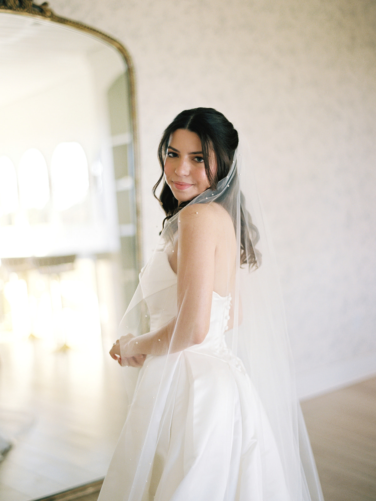 A woman in a white wedding dress and veil stands indoors, looking over her shoulder toward the camera.