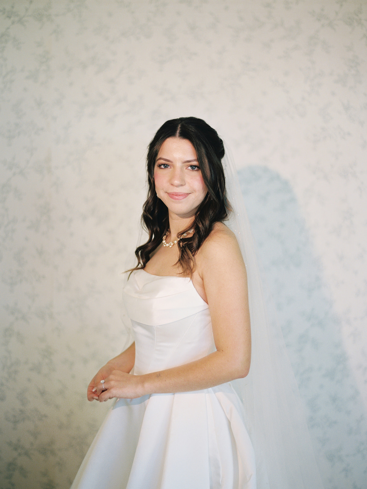 A woman in a white wedding dress and veil stands indoors, looking over her shoulder toward the camera.