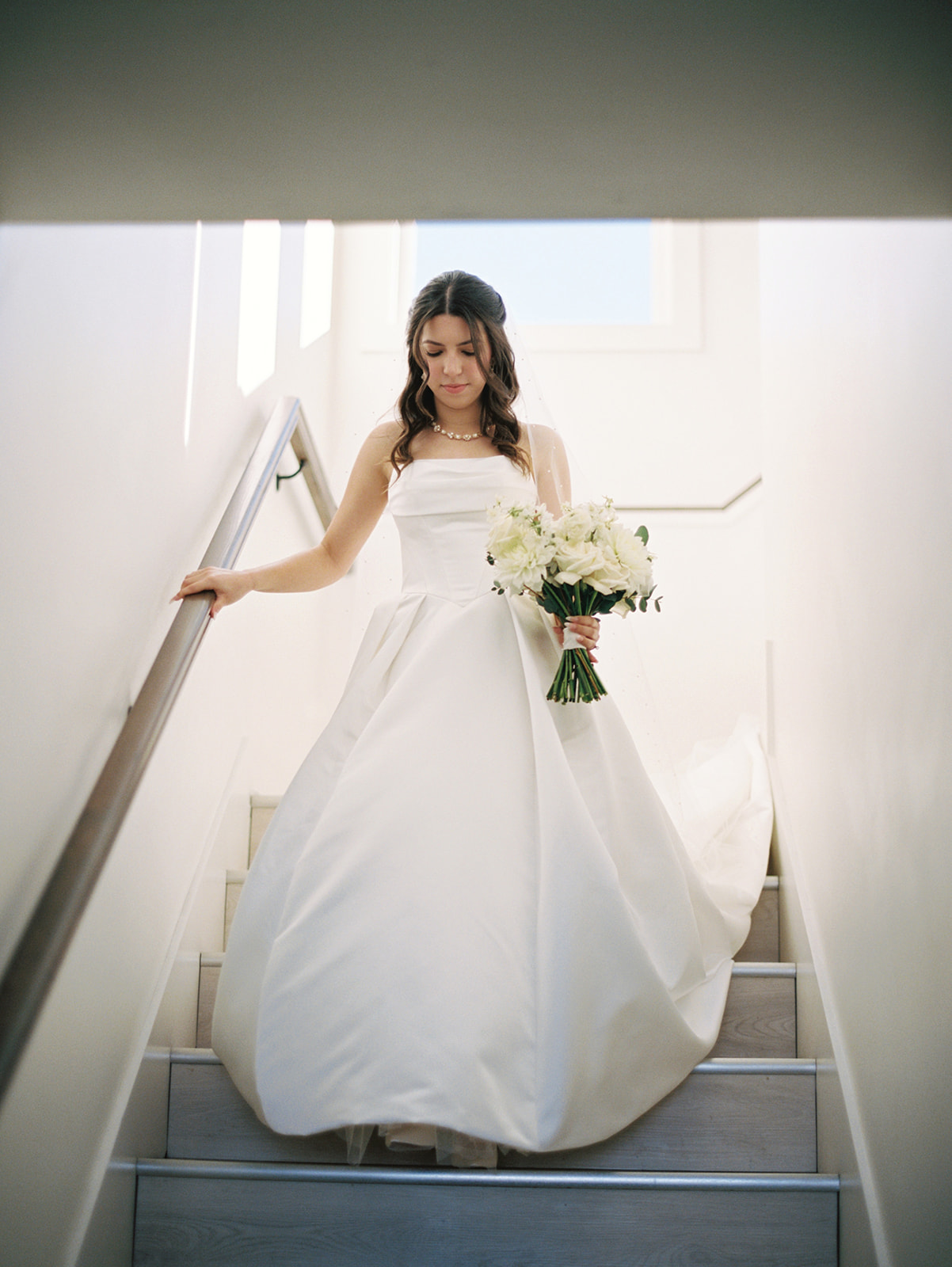 A bride in a white wedding dress descends a staircase, holding a bouquet of white flowers and gently touching the handrail.