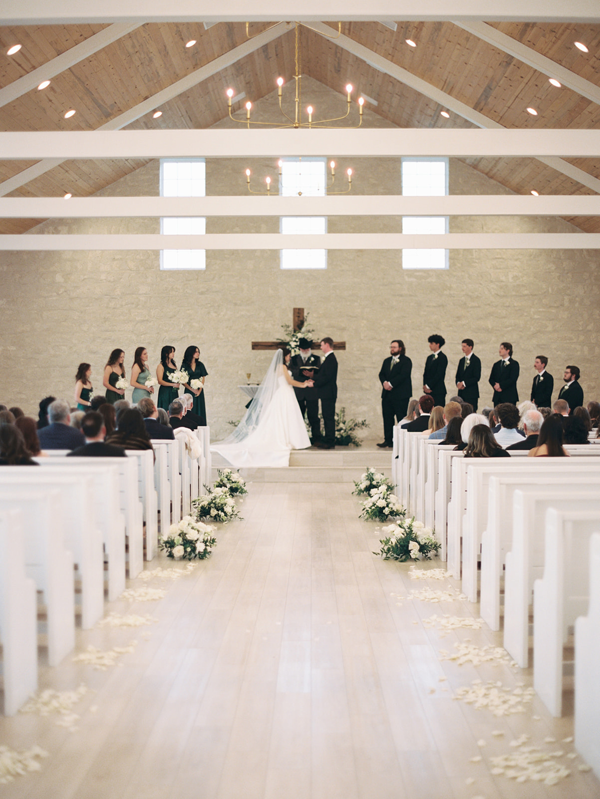 A bright chapel interior with wooden pews, floral arrangements along the aisle, and a large cross decorated with flowers at the front.