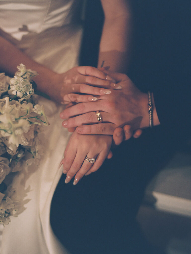 Two people in formal attire rest their hands together, showing wedding rings. One person holds a bouquet of white flowers.