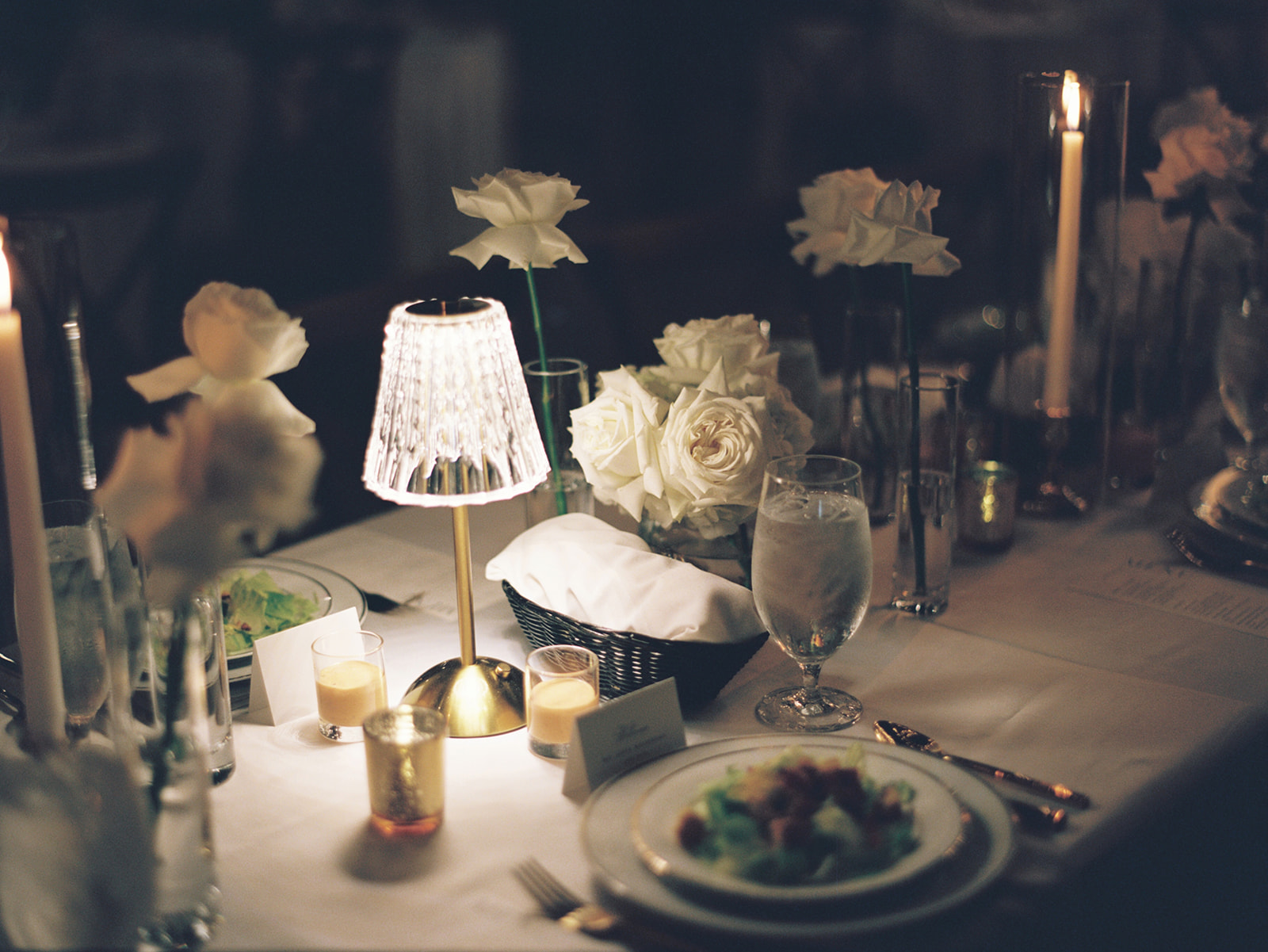A dimly lit dinner table is set with white roses, candles, a lamp, glassware, plates of food, and name cards.