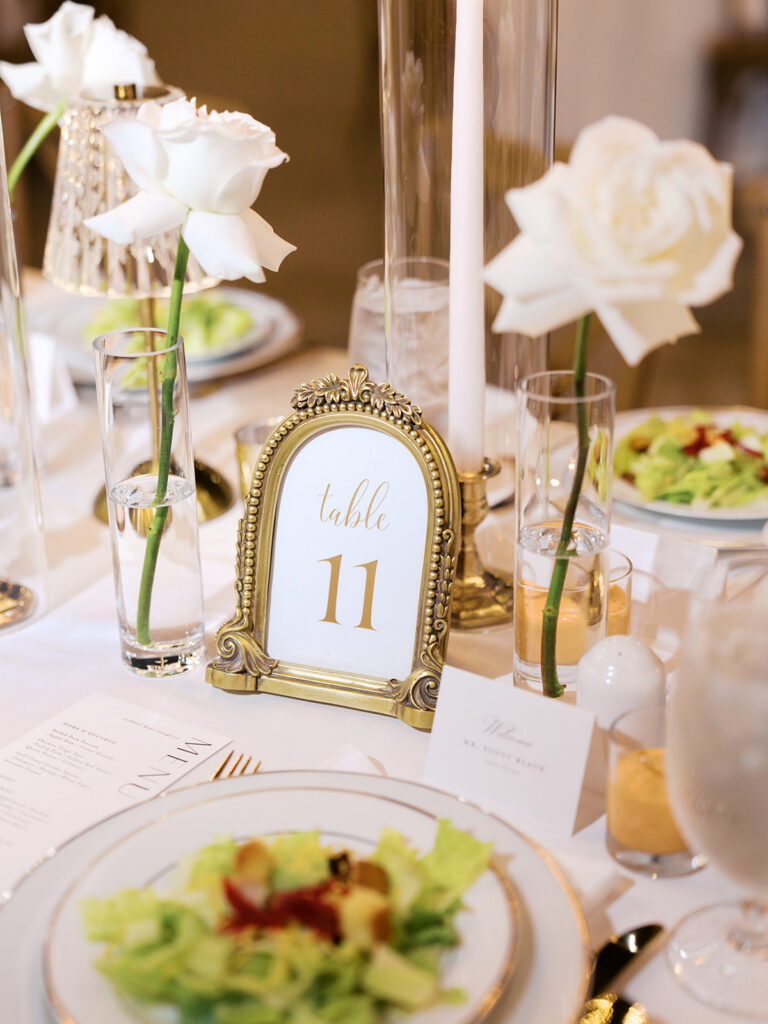 Elegant houston wedding decor with Long banquet table set with white tablecloths, plates of salad, wine glasses, and tall white candles in a bright, modern venue with large windows 