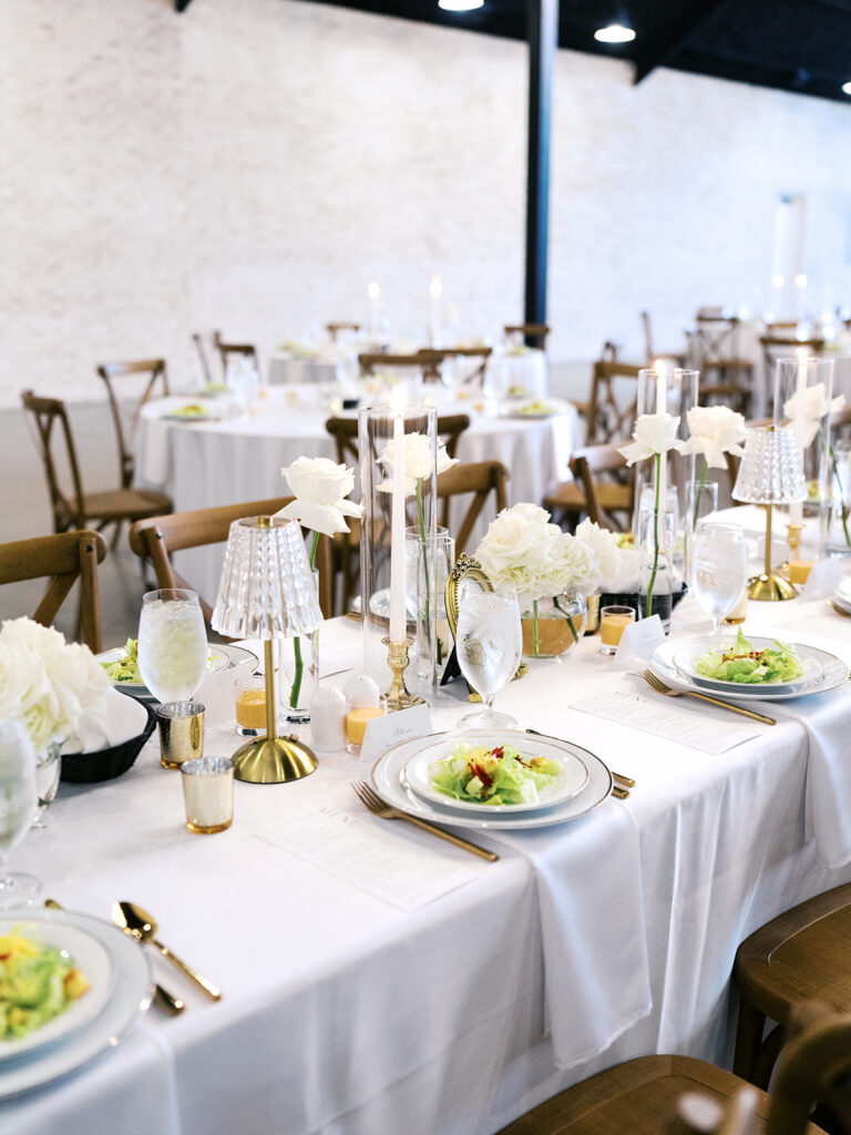 Elegant houston wedding decor with Long banquet table set with white tablecloths, plates of salad, wine glasses, and tall white candles in a bright, modern venue with large windows 